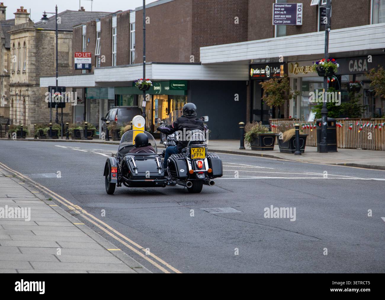 Vista posteriore di un motociclista con un sidecar che guida lungo una strada cittadina passando accanto ai negozi locali Melksham Wiltshire Foto Stock