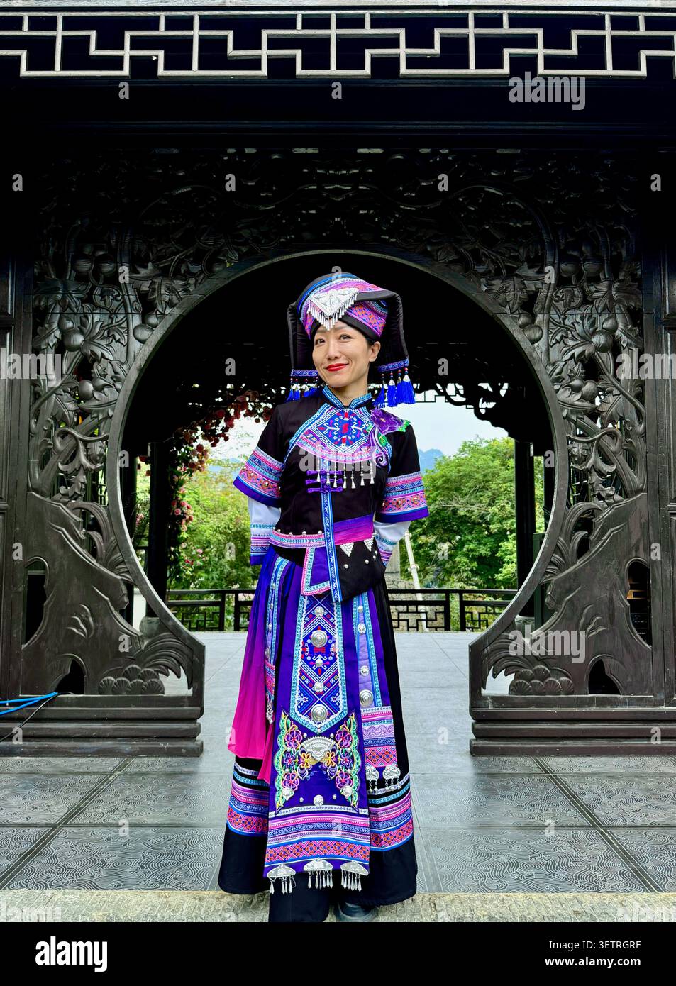 Donna con tradizionale costume ricamato da minoranze etniche incentrato su un cancello circolare in legno decorato intagliato, padiglione giardino patrimonio cinese, Guang - Immagine stock catturata con smartphone