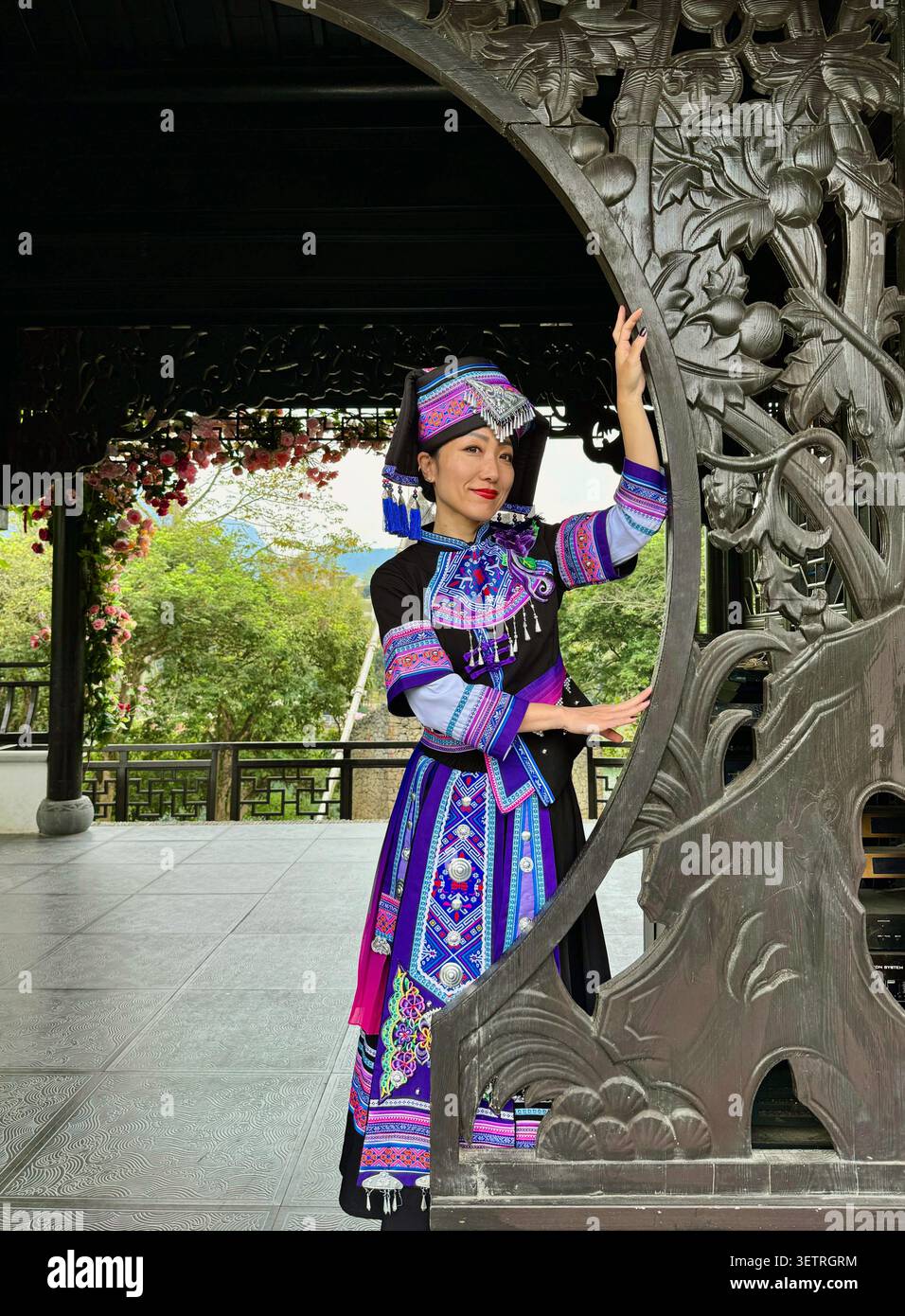Donna in costume tradizionale ricamato da minoranze etniche in posa accanto ad arco decorato con padiglione in legno intagliato con motivi di loto, giardino storico cinese, - Immagine stock catturata con smartphone