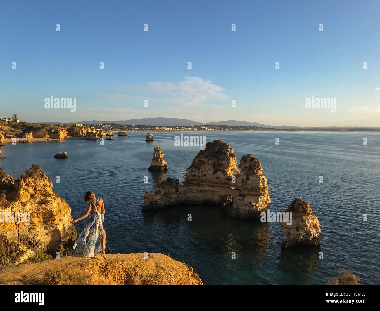 Una donna in un abito floreale fluente in piedi con grazia su un punto panoramico con vista sulla drammatica formazione rocciosa costiera. Scena di moda e stile di vita Foto Stock
