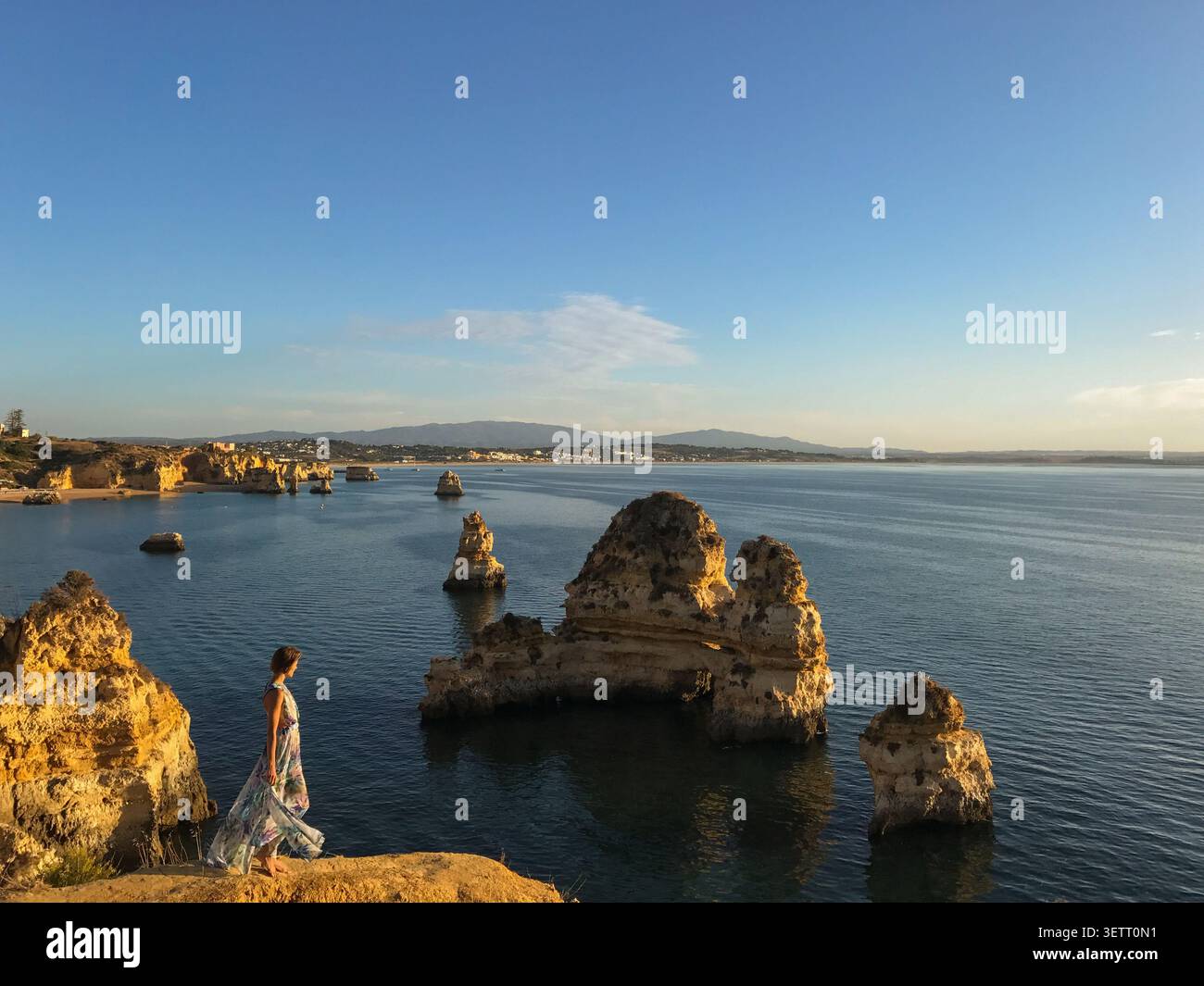 Una donna in un abito floreale fluente in piedi con grazia su un punto panoramico con vista sulla drammatica formazione rocciosa costiera. Scena di moda e stile di vita Foto Stock