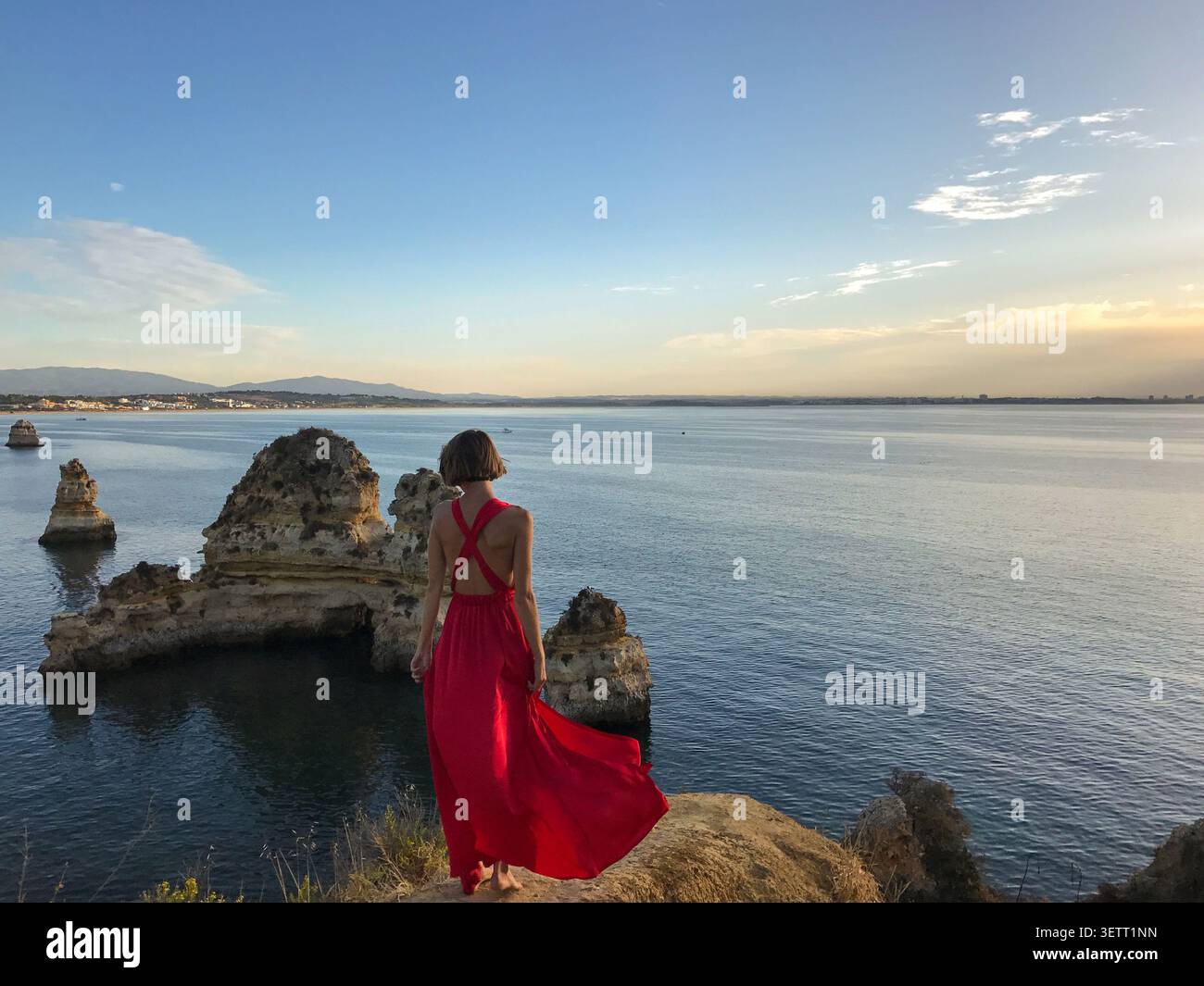 Una donna in un abito rosso fluente in posa elegante su un punto panoramico che si affaccia sulle drammatiche formazioni rocciose costiere. Scattato di spalle. Foto Stock