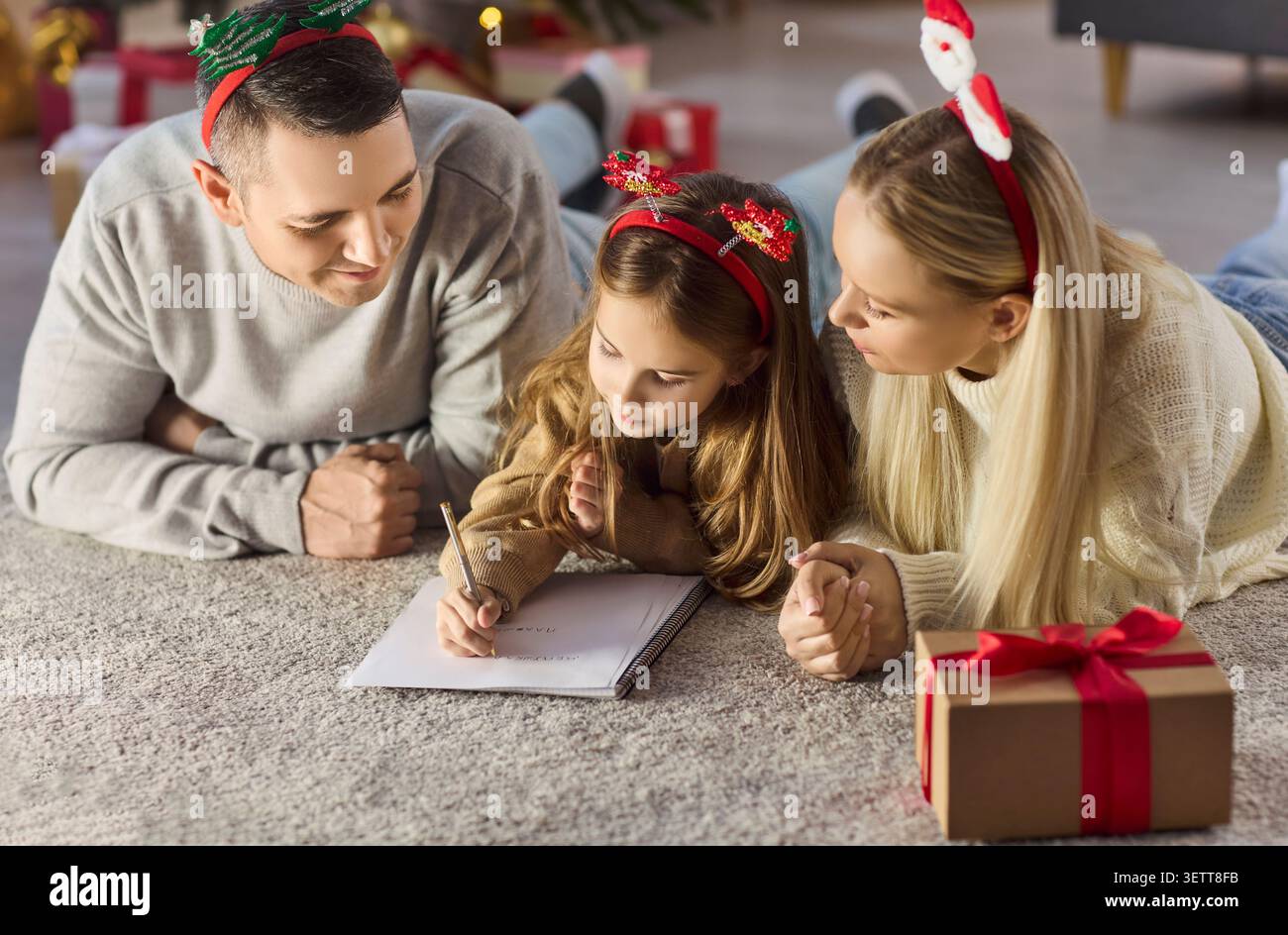 Bambino che scrive la lettera di Natale con i genitori sul pavimento, vicino al regalo e all'albero a casa Foto Stock