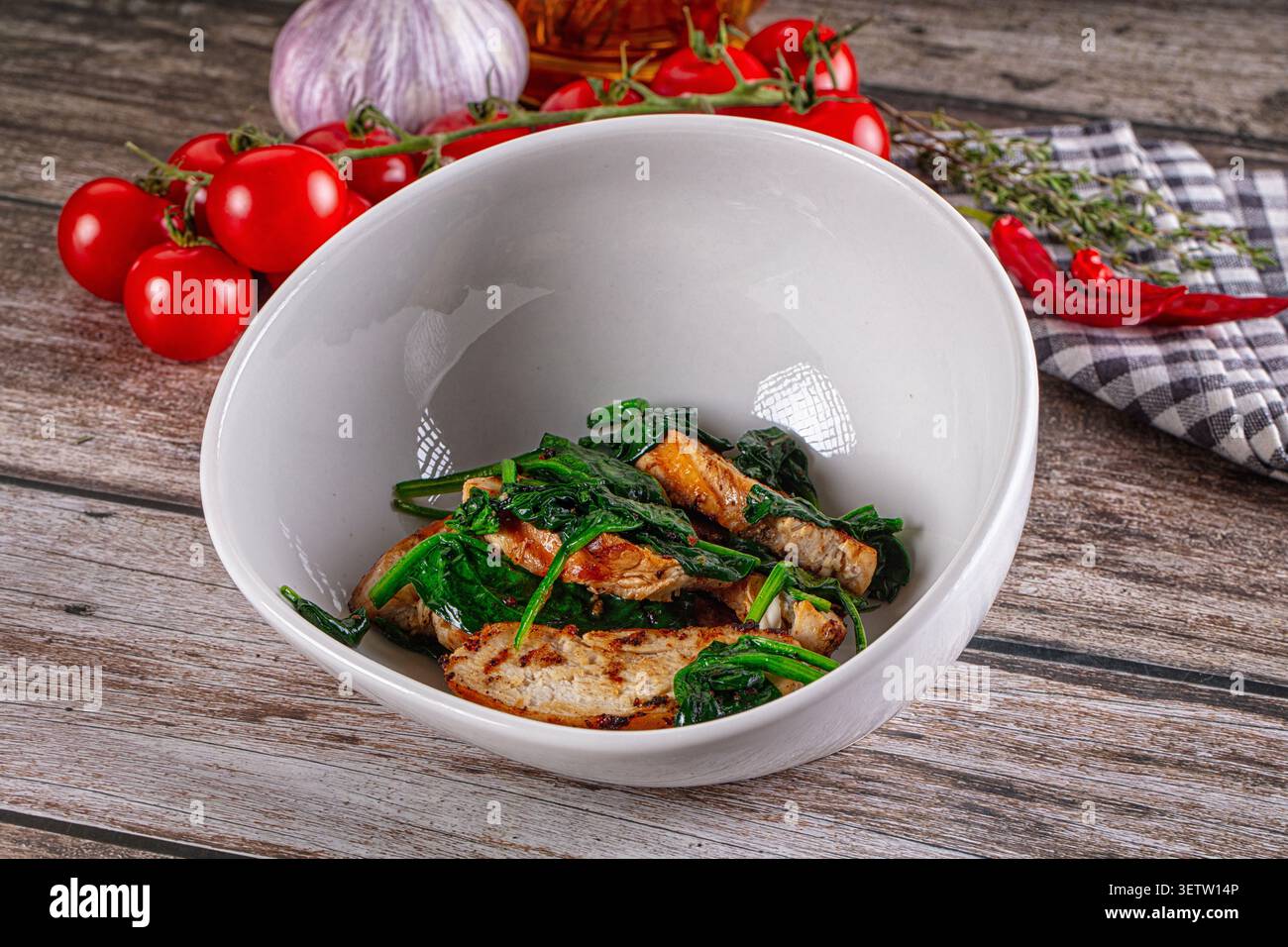 Fette di pollo alla griglia con spinaci selvatici in un recipiente di ceramica bianca. Superficie rustica in legno con pomodori ciliegini, aglio, peperoncino rosso, timo nella schiena Foto Stock
