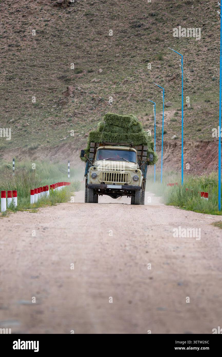 Un camion d'epoca dell'epoca sovietica di colore giallo, carico di balle di fieno verde, percorre una tortuosa strada sterrata ai piedi delle colline del Kirghizistan Foto Stock