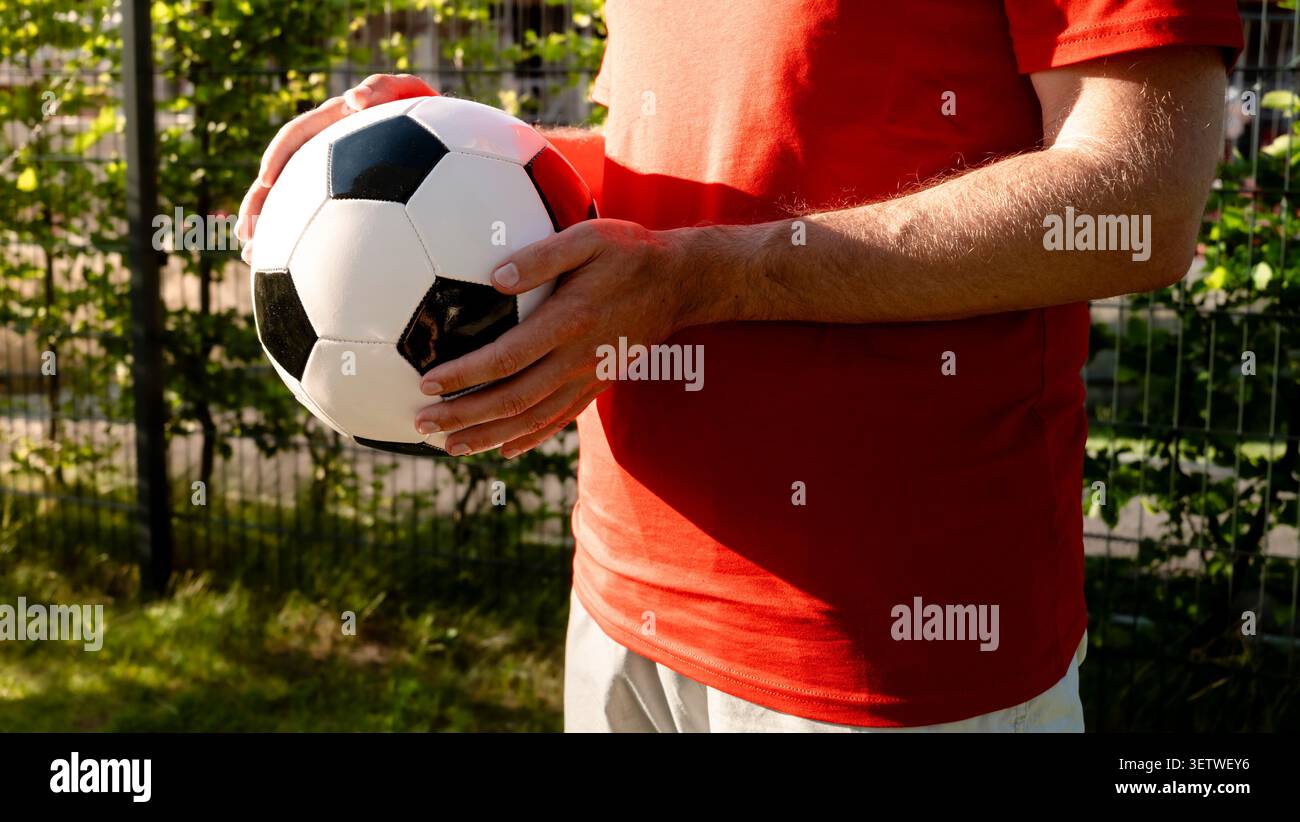 Persona con una maglietta rossa che regge un pallone da calcio bianco e nero, esprimendo lo spirito del calcio universale attraverso i colori classici. Foto Stock