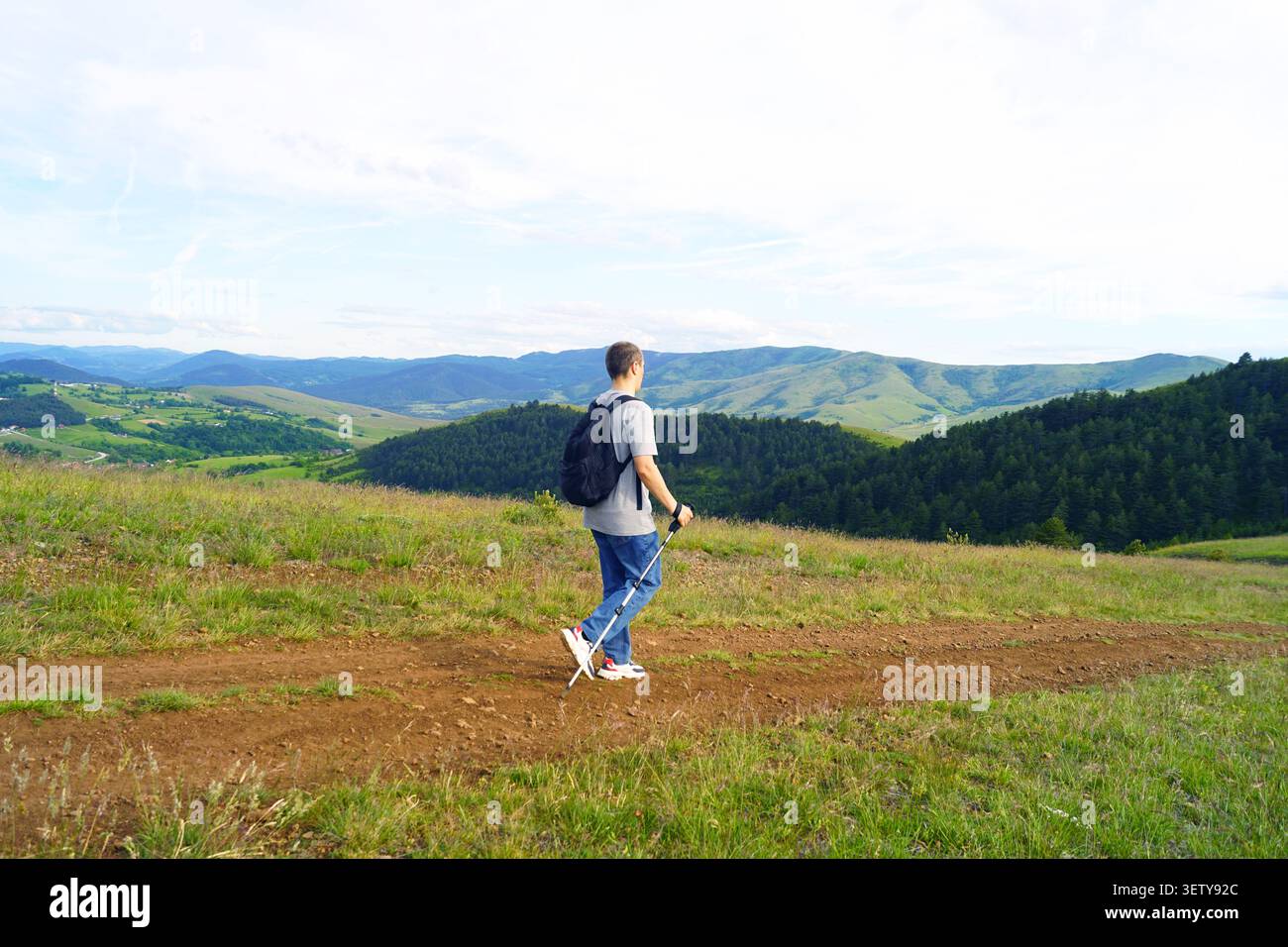 Escursionista che segue un percorso di campagna senza segnaletica verso il verde rigoglioso con pali da trekking. Mobilità indipendente e rifugio tranquillo dalla routine urbana. Foto Stock