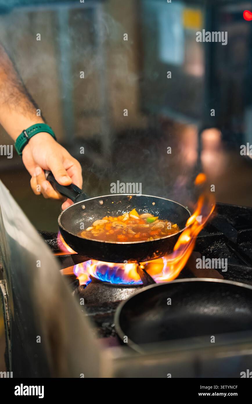 Lo chef tiene una padella nera sopra una fiamma di gas ruggente nella cucina professionale Foto Stock