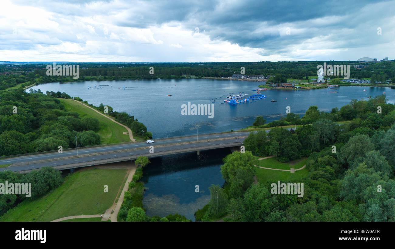 Vista aerea del lago Willen con un parco acquatico con ostacoli gonfiabili, un ponte e lussureggianti alberi verdi sotto un cielo nuvoloso a Milton Keynes, Engla Foto Stock
