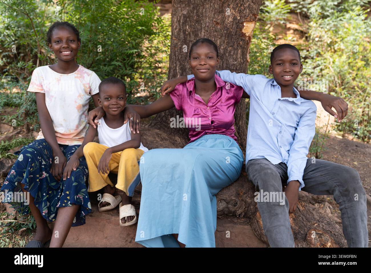 Fratelli africani, due sorelle e due fratelli siedono insieme sotto un albero con le braccia intorno alle spalle, e i loro sorrisi gioiosi riflettono Foto Stock