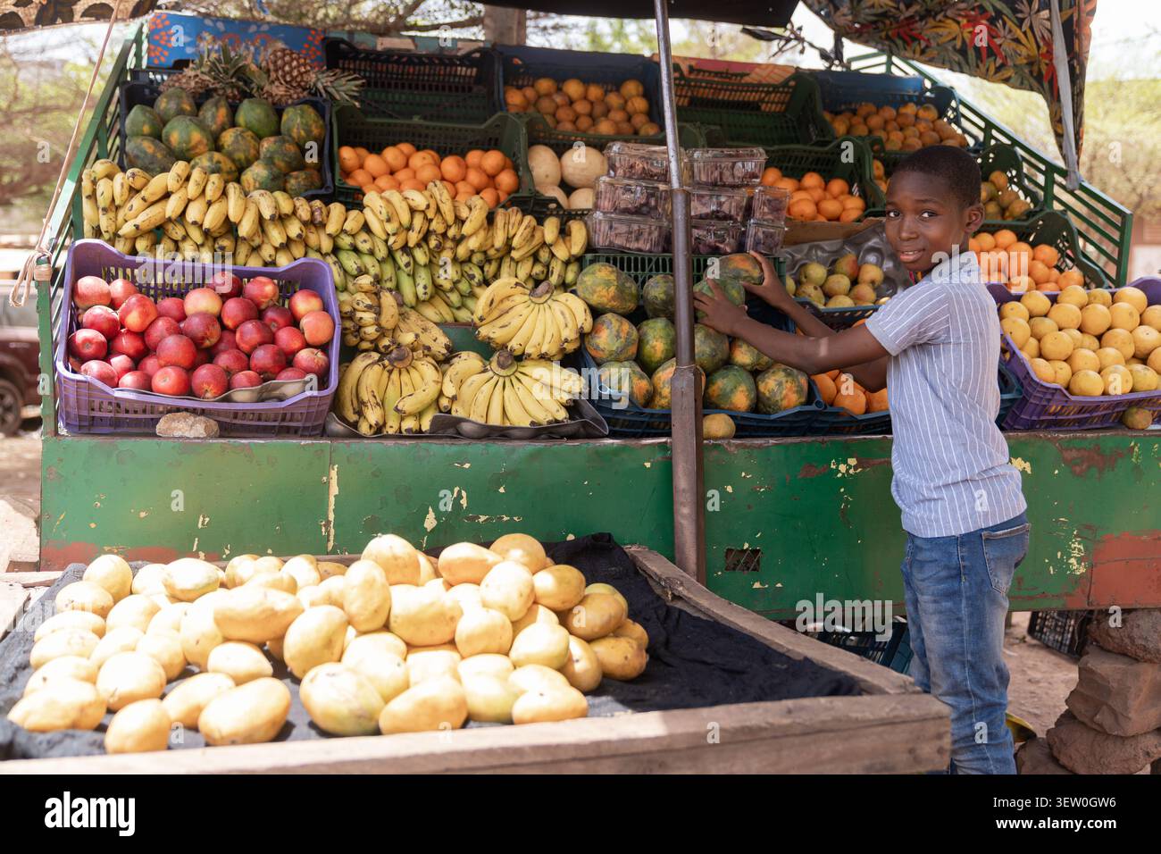 Un ragazzo africano si trova in una bancarella di frutta colorata, sorridendo mentre regge una papaia tra mucchi di banane, arance e manghi. Foto Stock