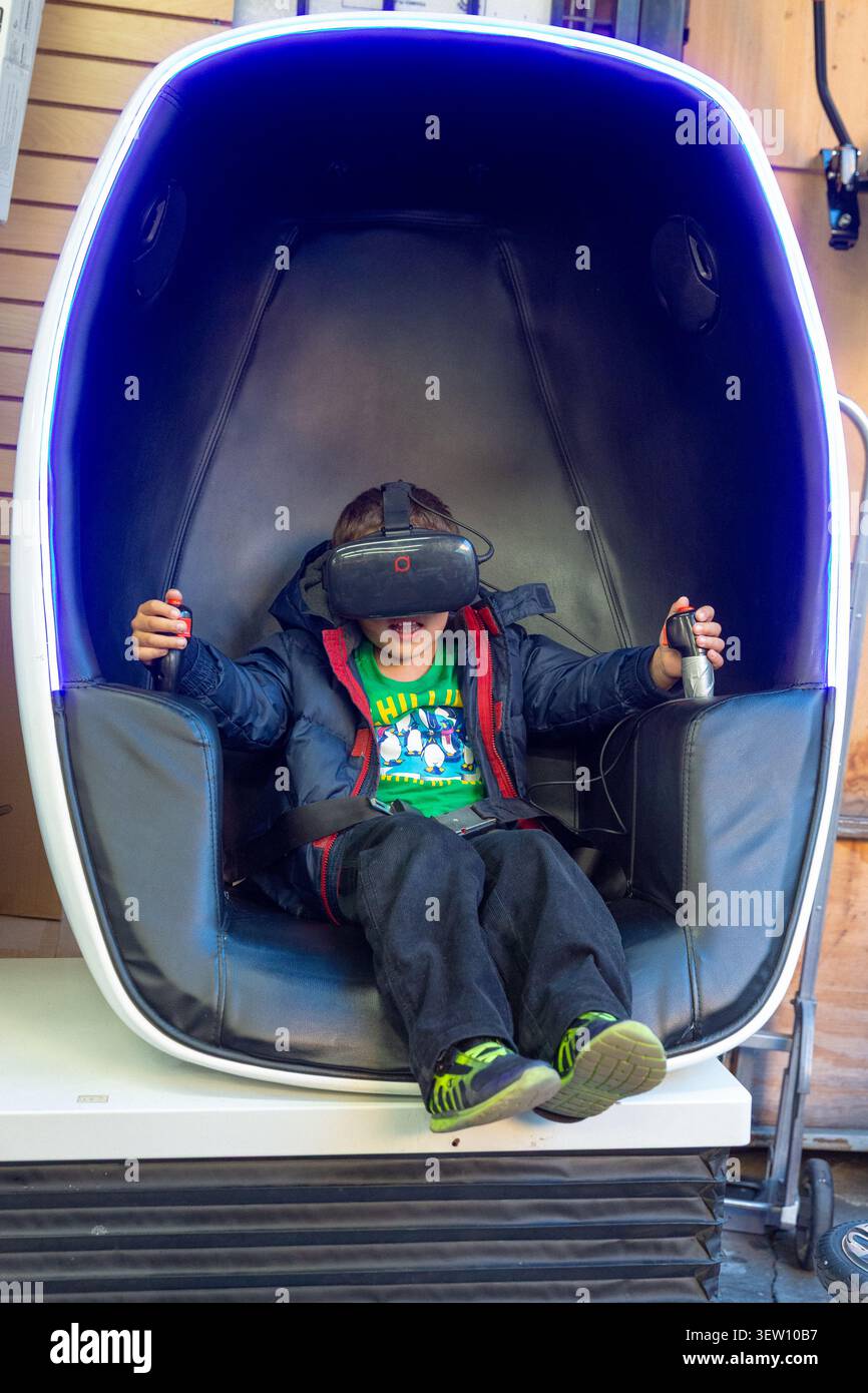 Un bambino in una poltrona VR (simulatore di realtà virtuale immersivo), indossa un visore, mani sui joystick, in un negozio nel centro di Los Angeles. Foto Stock