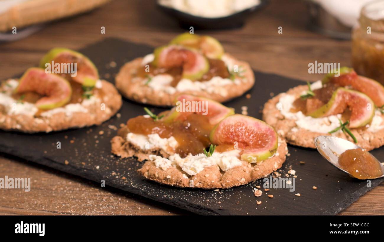 Una selezione di pani croccanti artigianali di grano intero serviti su un tabellone di ardesia nera, ciascuno ricoperto da uno strato di formaggio cremoso, confettura di fichi dolci e fi fresco Foto Stock