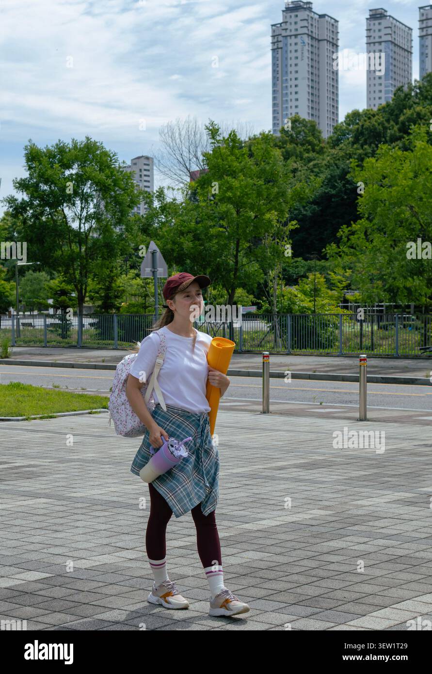 Giovane donna che cammina per una lezione di yoga all'aperto portando con sé una stuoia di esercizio nell'Urban Park Foto Stock