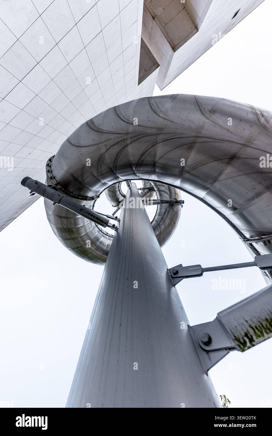 Dettagli architettonici che mostrano una scultura dinamica a spirale in acciaio inox che si avvicina verso l'alto verso un edificio geometrico moderno, con un supporto centrale p Foto Stock