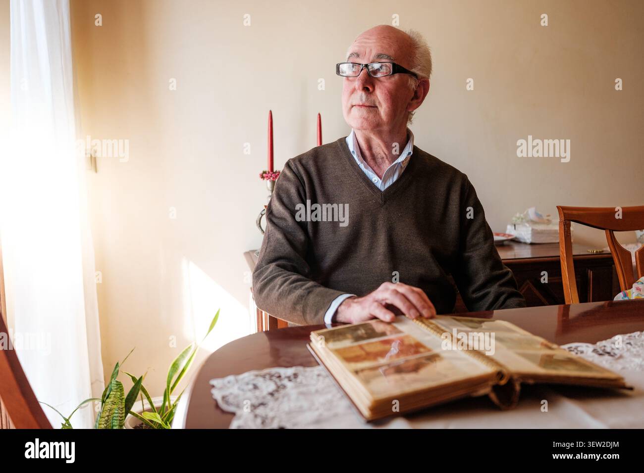 Uomo anziano seduto a tavola, a riflettere sulla nostalgia mentre tiene un album fotografico aperto Foto Stock