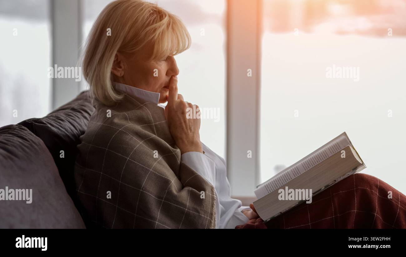 Donna anziana premurosa seduta comodamente su un divano, coperta da una coperta a quadri, che contempla mentre legge un libro alla luce della finestra, godendo di un Foto Stock