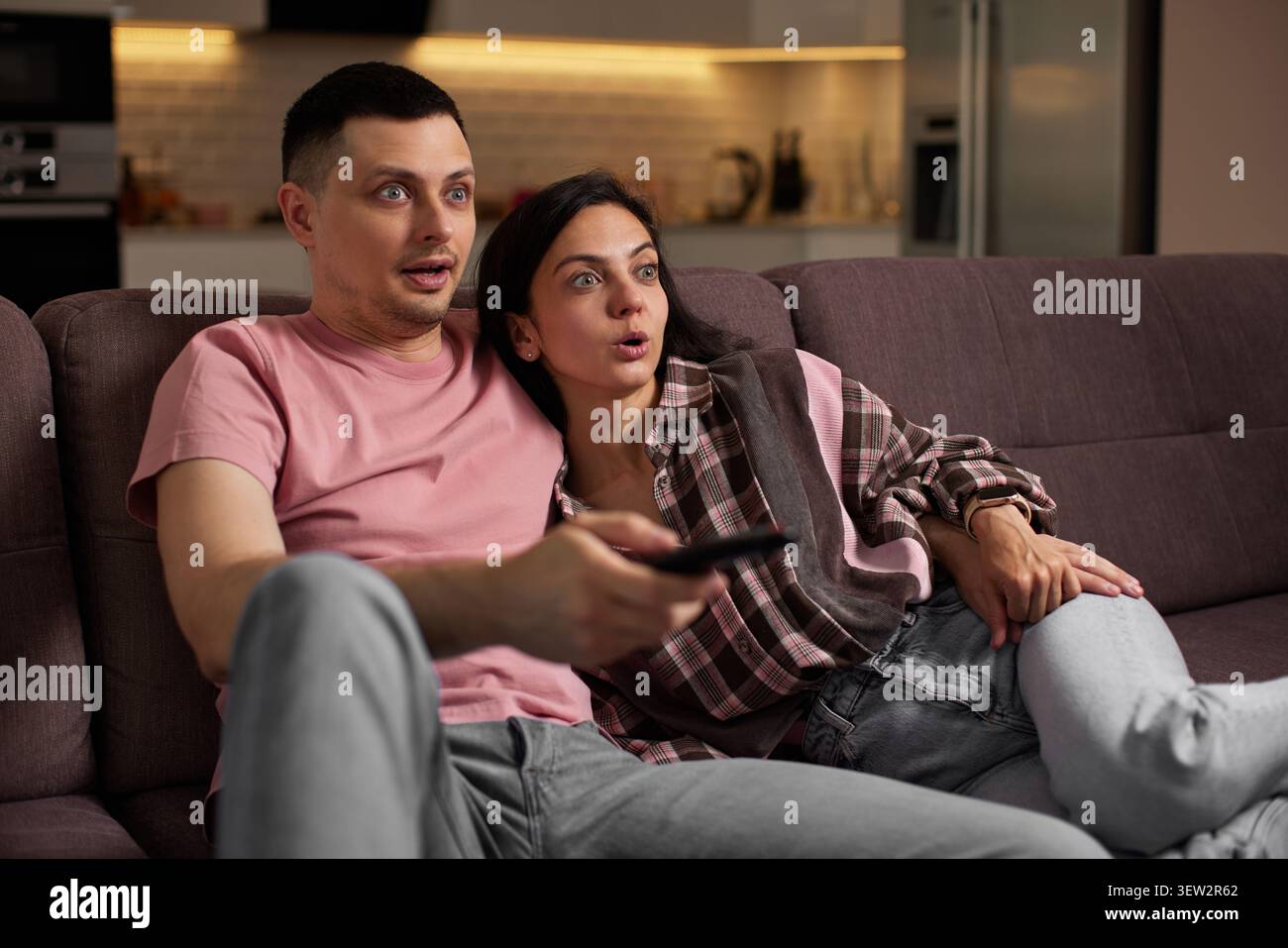 Una giovane coppia seduta su un divano nel soggiorno, sembra sorpresa, stupita e scioccata mentre guarda la televisione Foto Stock
