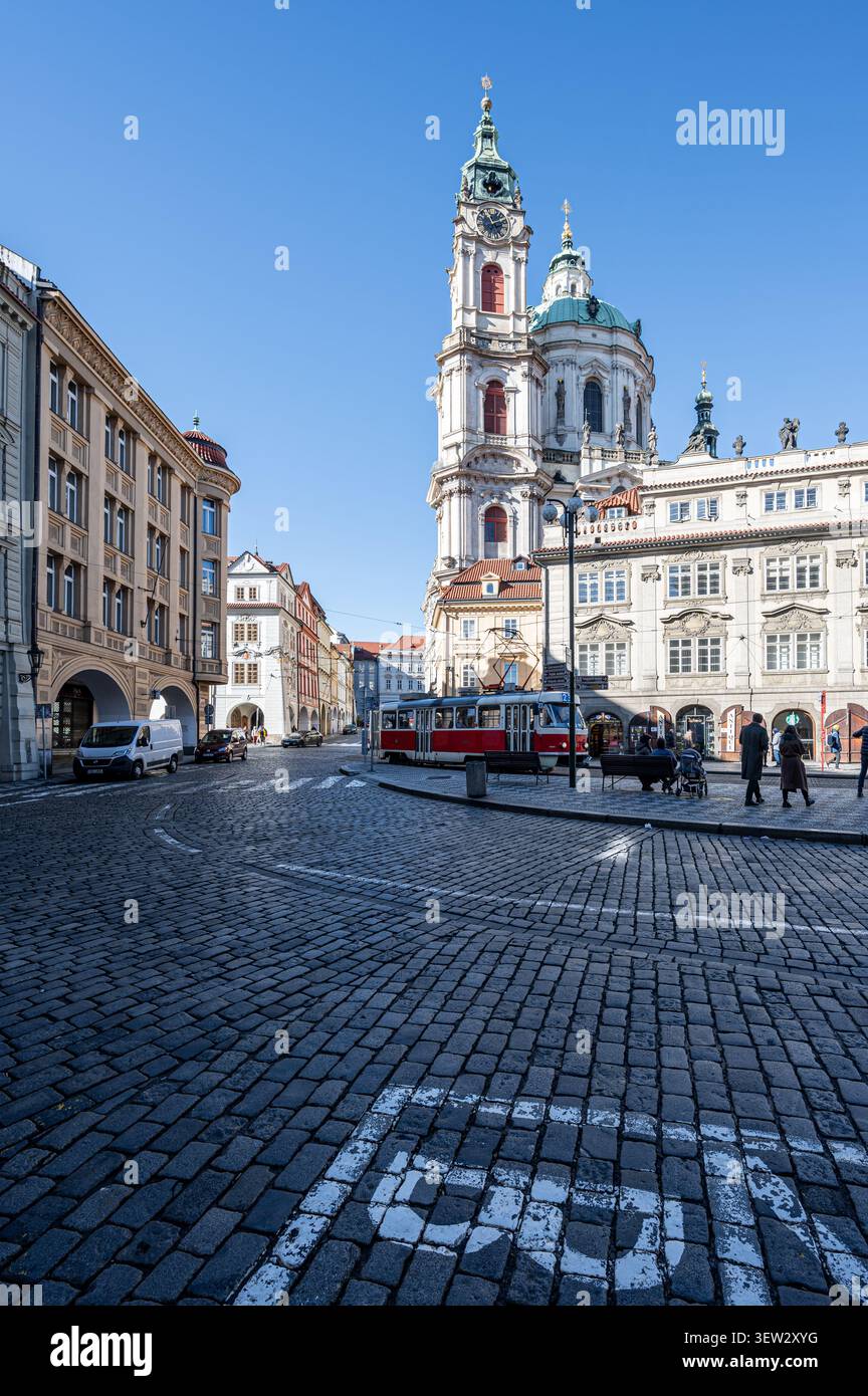Un tram rosso tradizionale passa attraverso una piazza lastricata di ciottoli davanti all'architettura barocca della Chiesa di San Nicola nel quartiere di Mala Strana di Foto Stock