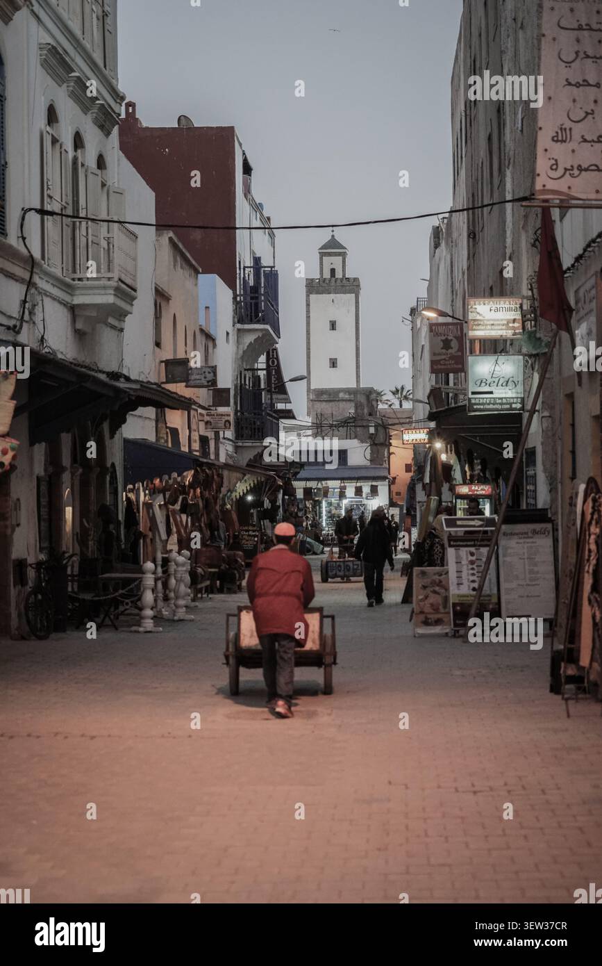 Essaouira, Marocco - 5 dicembre 2017: Un uomo con una giacca rossa spinge un carro lungo una stretta strada acciottolata nella Medina di Essaouira al crepuscolo, con una p Foto Stock
