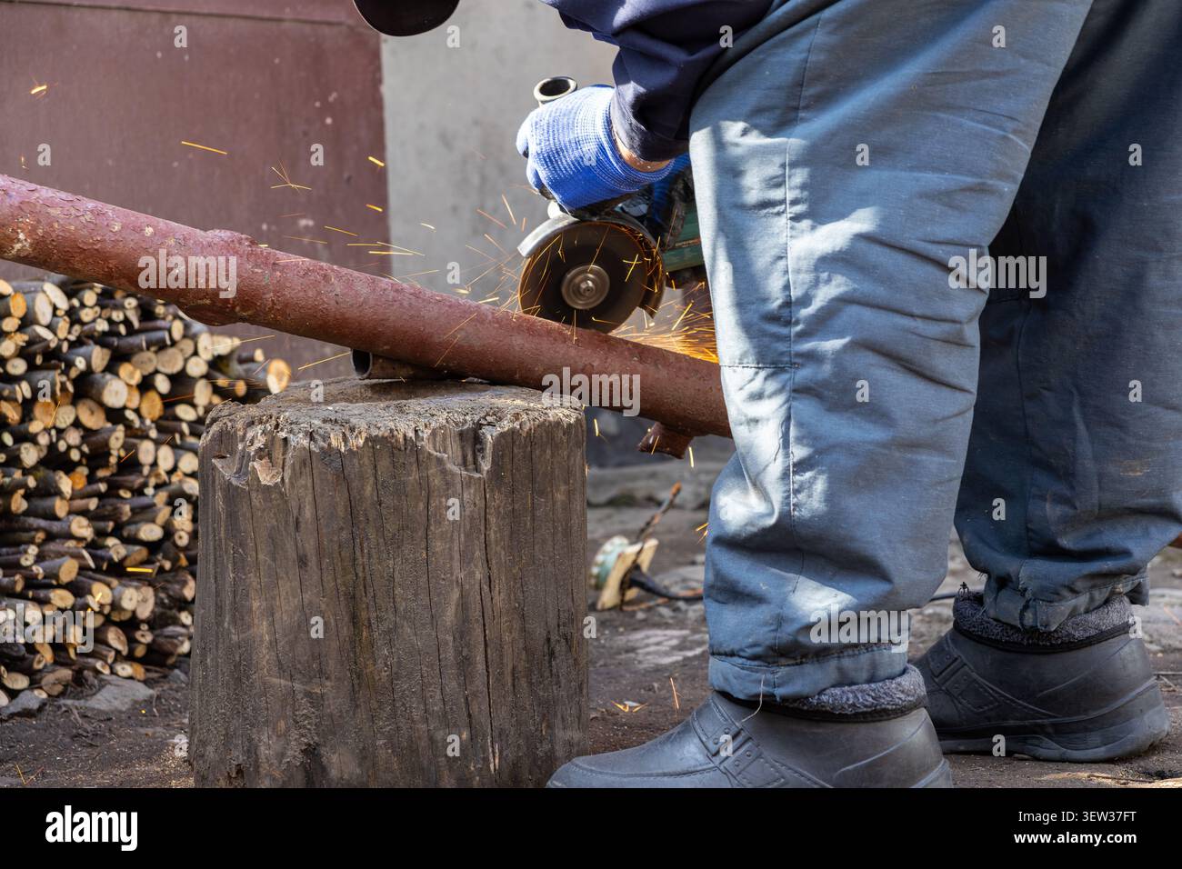 Il tecnico con guanti protettivi blu aziona un utensile elettrico per tagliare un tubo intemprato, creando scintille volanti che enfatizzano l'intensità di Foto Stock