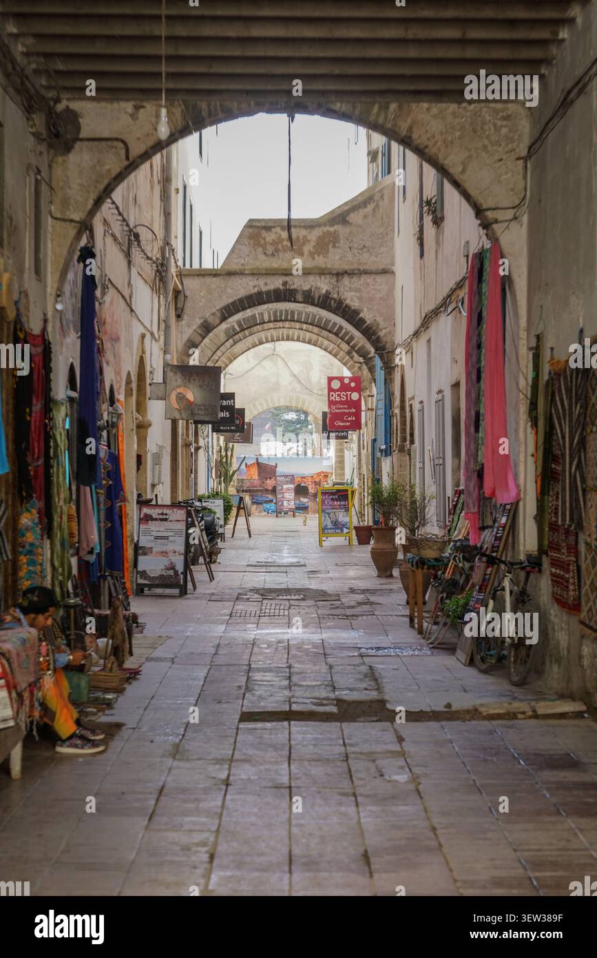 Essaouira, Marocco - 5 dicembre 2017: Una lunga e stretta strada nella medina di Essaouira è vista attraverso una serie di archi in pietra, fiancheggiati da negozi Foto Stock
