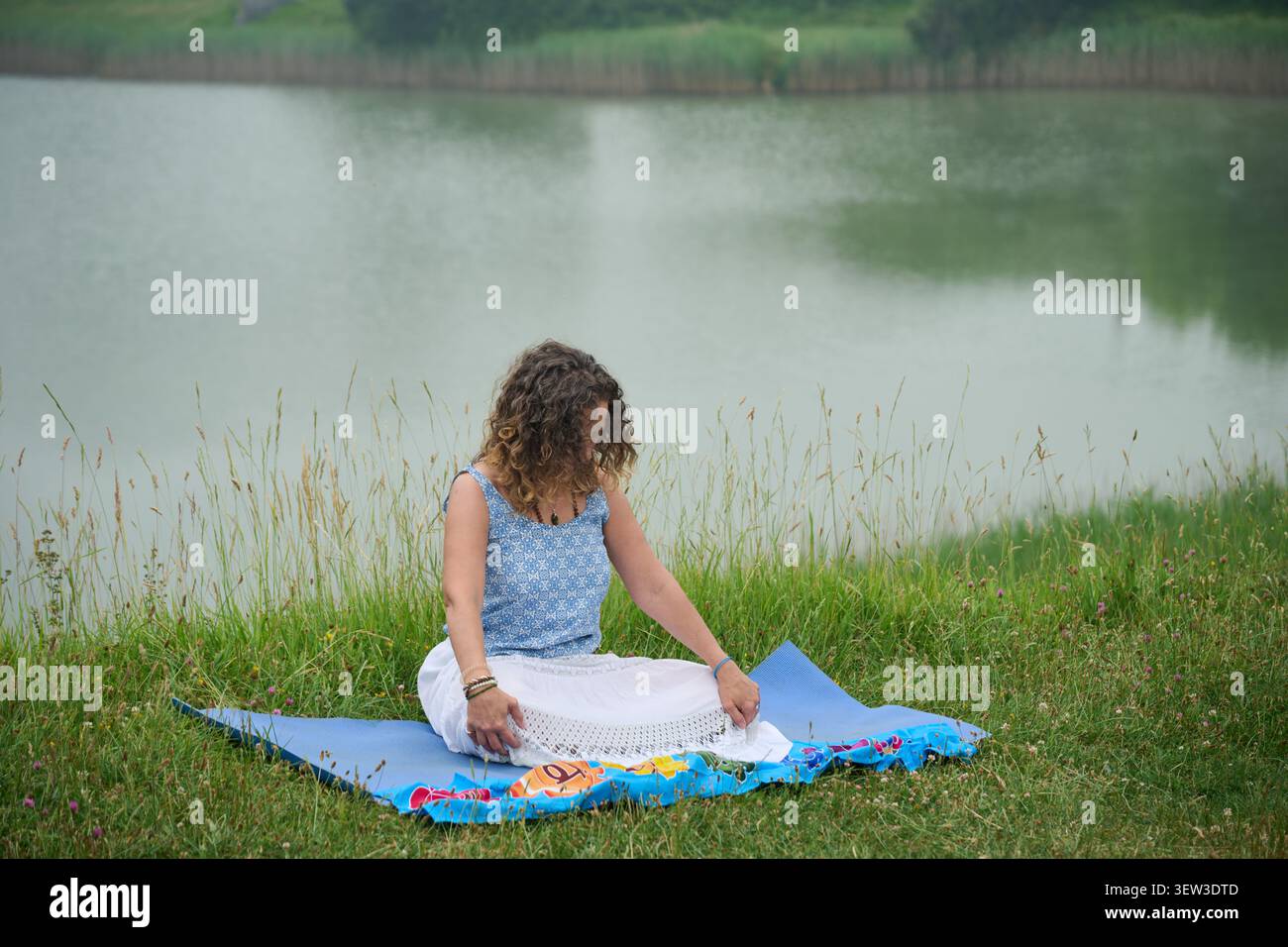 Donna seduta su un tappeto vicino al lago in un campo erboso, godendo di un momento di pace e tranquillità, praticando consapevolezza e cura di sé in natura Foto Stock