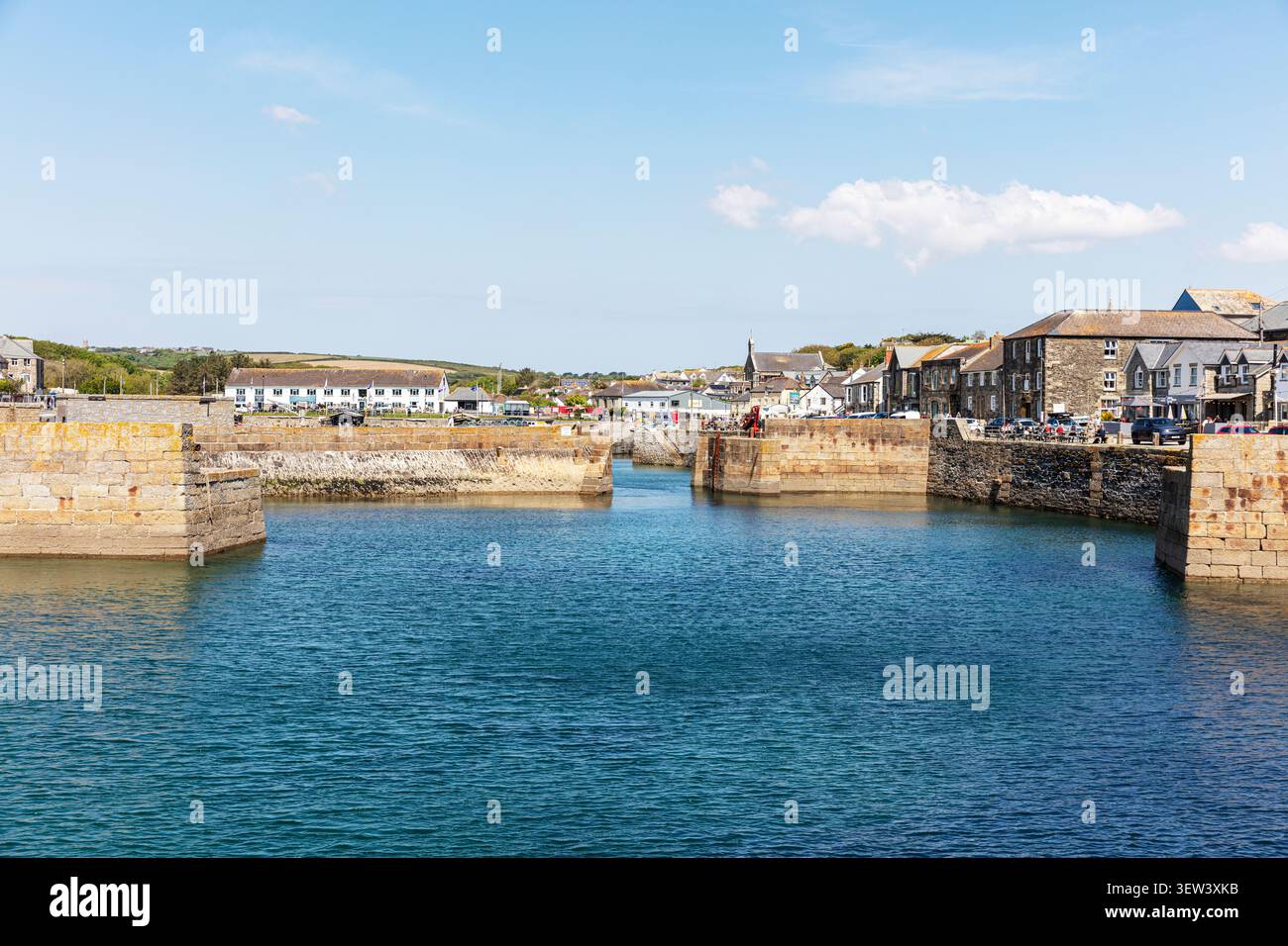 Ingresso al porto di Porthleven, Cornovaglia, con acque blu calme delimitate da mura in pietra del porto. Gli edifici costieri e le attività costiere costeggiano il lungomare e Foto Stock
