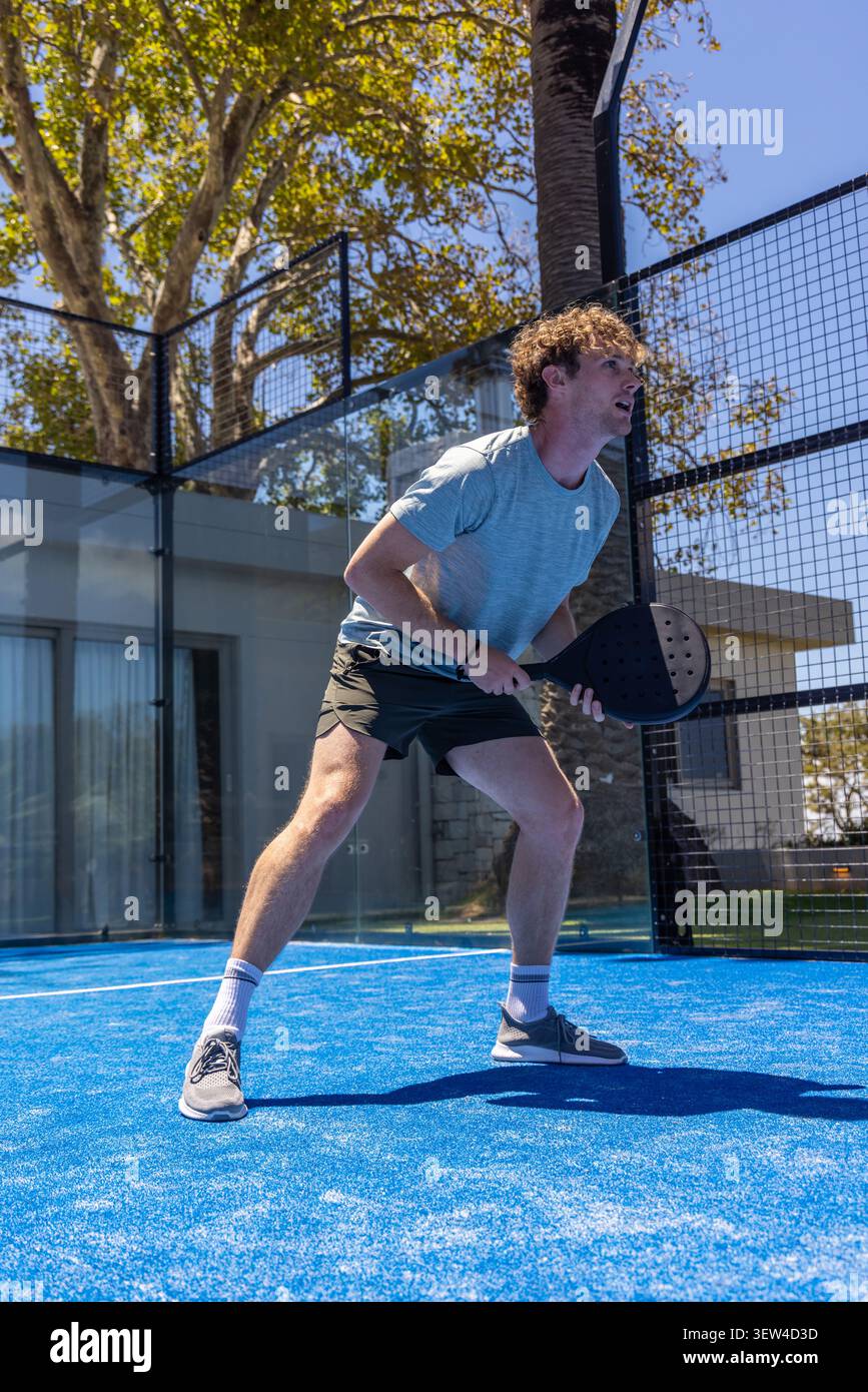 Uomo in abbigliamento sportivo in piedi su un campo in padel blu brillante che regge la racchetta e guarda in alto Foto Stock