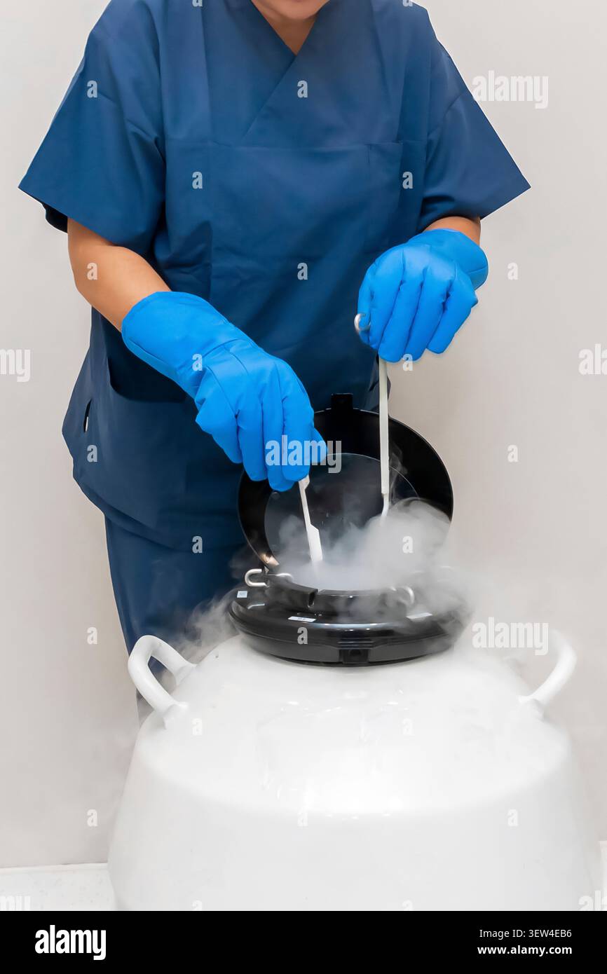 Embriologo maschio in camici blu e guanti criogenici che lavora con un serbatoio di azoto liquido. Conservazione degli embrioni mediante congelamento in un moderno laboratorio di clinica IVF. Foto Stock