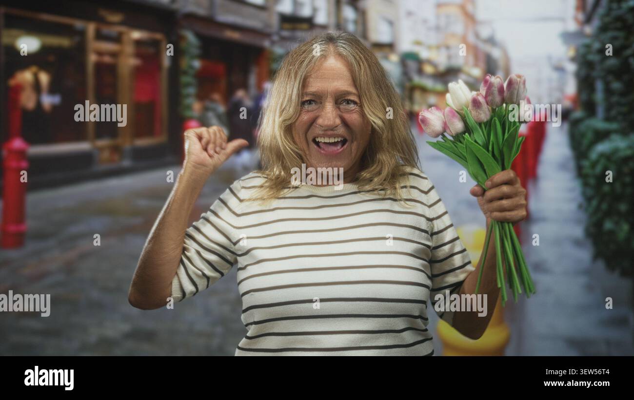 Donna che tiene i tulipani e alza il pugno mentre ride, gli occhi chiusi, indossa una camicia a righe in una trafficata strada dello shopping con bollitori rossi e magazzini Foto Stock