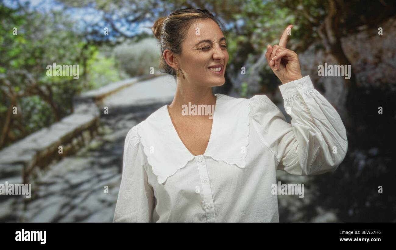 La donna punta il dito indice verso l'alto sul sentiero della foresta, gli occhi chiusi e sorridenti in camicetta bianca; momento di gioia. Foto Stock
