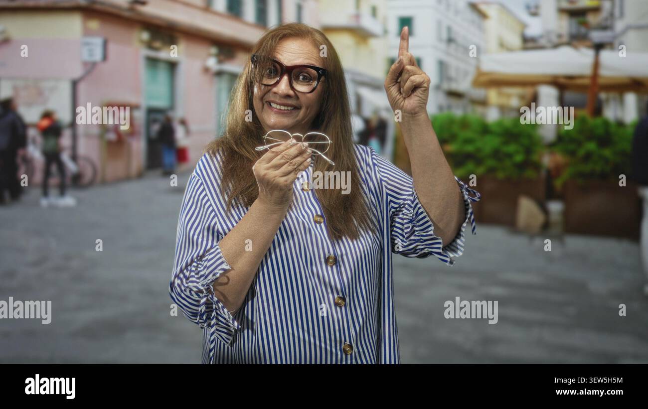 Donna anziana che sorride, tiene gli occhiali e punta il dito su una strada pedonale vicino a ombrelloni e piantagioni vicino a edifici pastello; disco gioioso Foto Stock