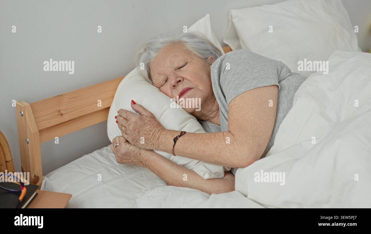 Una donna anziana abbraccia un cuscino mentre dorme su un letto in una camera da letto, distesa sul fianco con gli occhi chiusi e le mani che stringono il cuscino; comfort serenità. Foto Stock