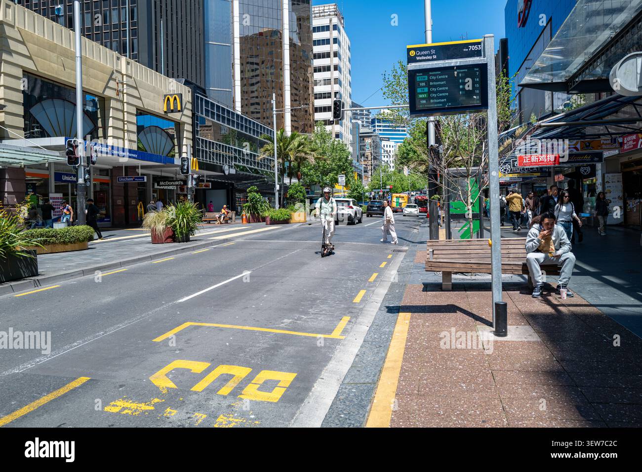 Un pendolare su uno scooter elettrico passa davanti a un cartello digitale di transito e a una corsia bus su Queen Street di Auckland, illustrando le tendenze della micro-mobilità urbana. Foto Stock