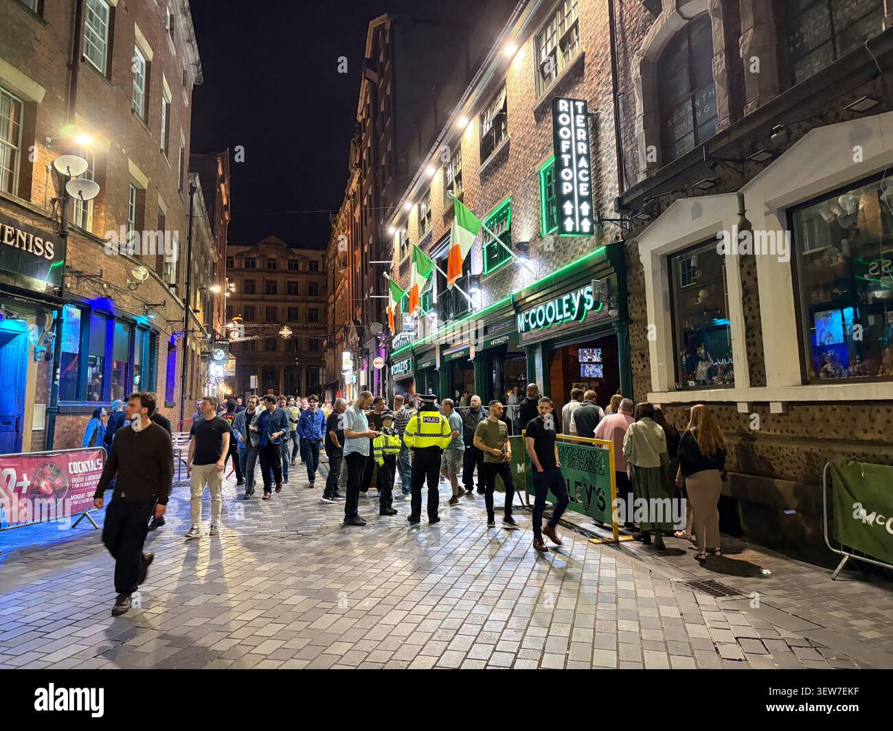 I pedoni si riuniscono in una strada lastricata fuori dal bar McCooly's a Liverpool, in Inghilterra, durante la notte con cartelli illuminati. Foto Stock