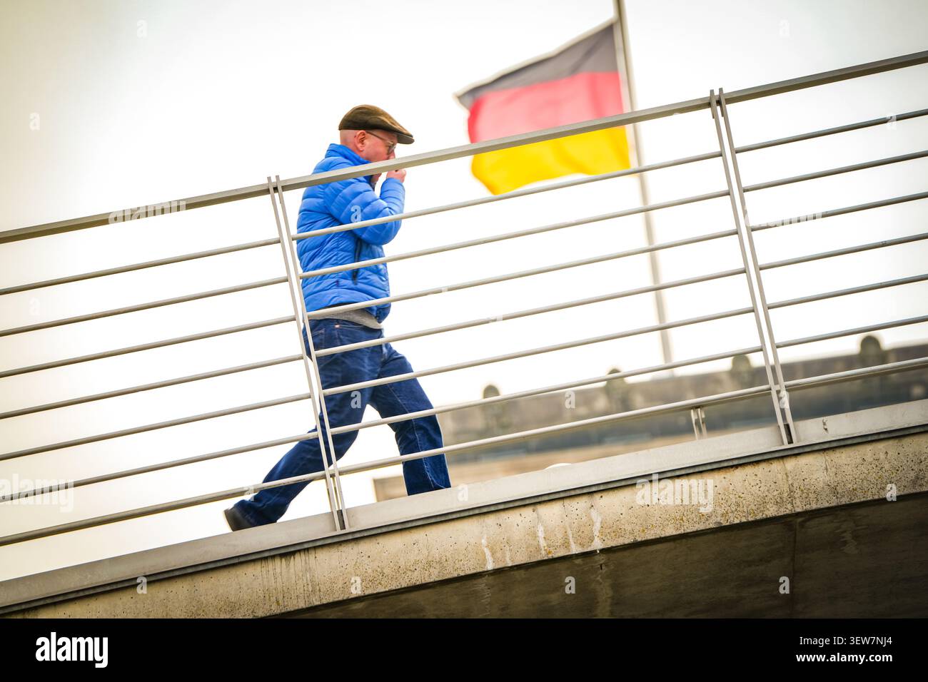 Figura solitaria in giacca blu che passa accanto a ringhiere e a una bandiera tedesca sventolata dal vento, Berlino Foto Stock