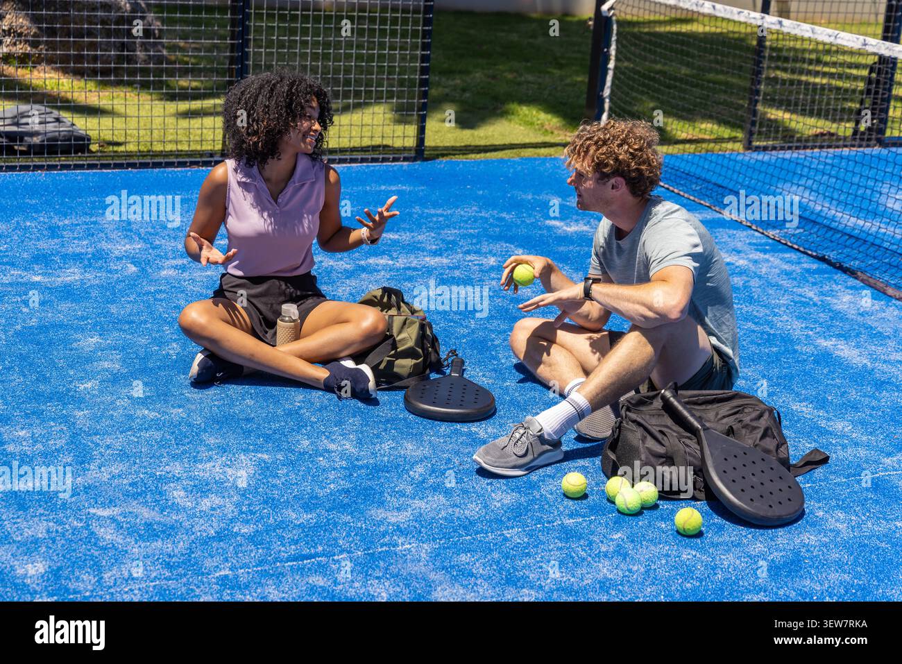 Diversi amici seduti a gambe incrociate su un campo blu con abbigliamento sportivo, pagaie e palle da tennis Foto Stock
