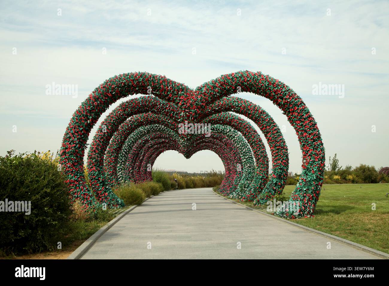 Un liscio sentiero pavimentato conduce attraverso un romantico tunnel di archi floreali a forma di cuore a più strati. Ogni arco è densamente ricoperto di tallone artificiale Foto Stock