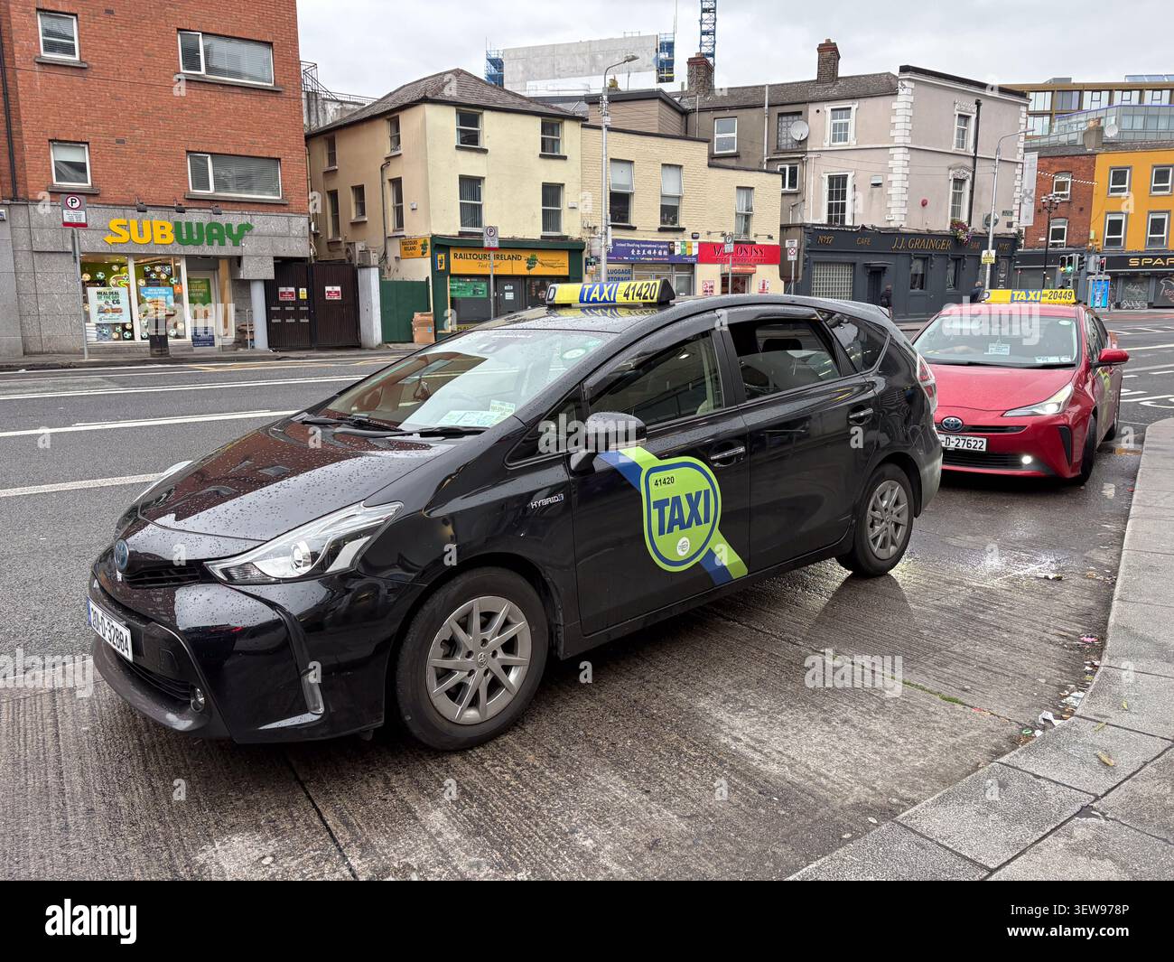 Due moderni taxi ibridi parcheggiati in una strada cittadina di Dublino, Irlanda, con edifici commerciali e vetrine sullo sfondo. Foto Stock