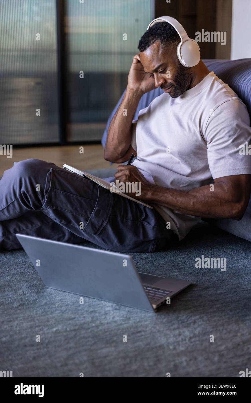 Uomo africano maturo seduto su un tappeto accanto al divano a casa, scrivendo su un notebook con laptop e cuffie Foto Stock