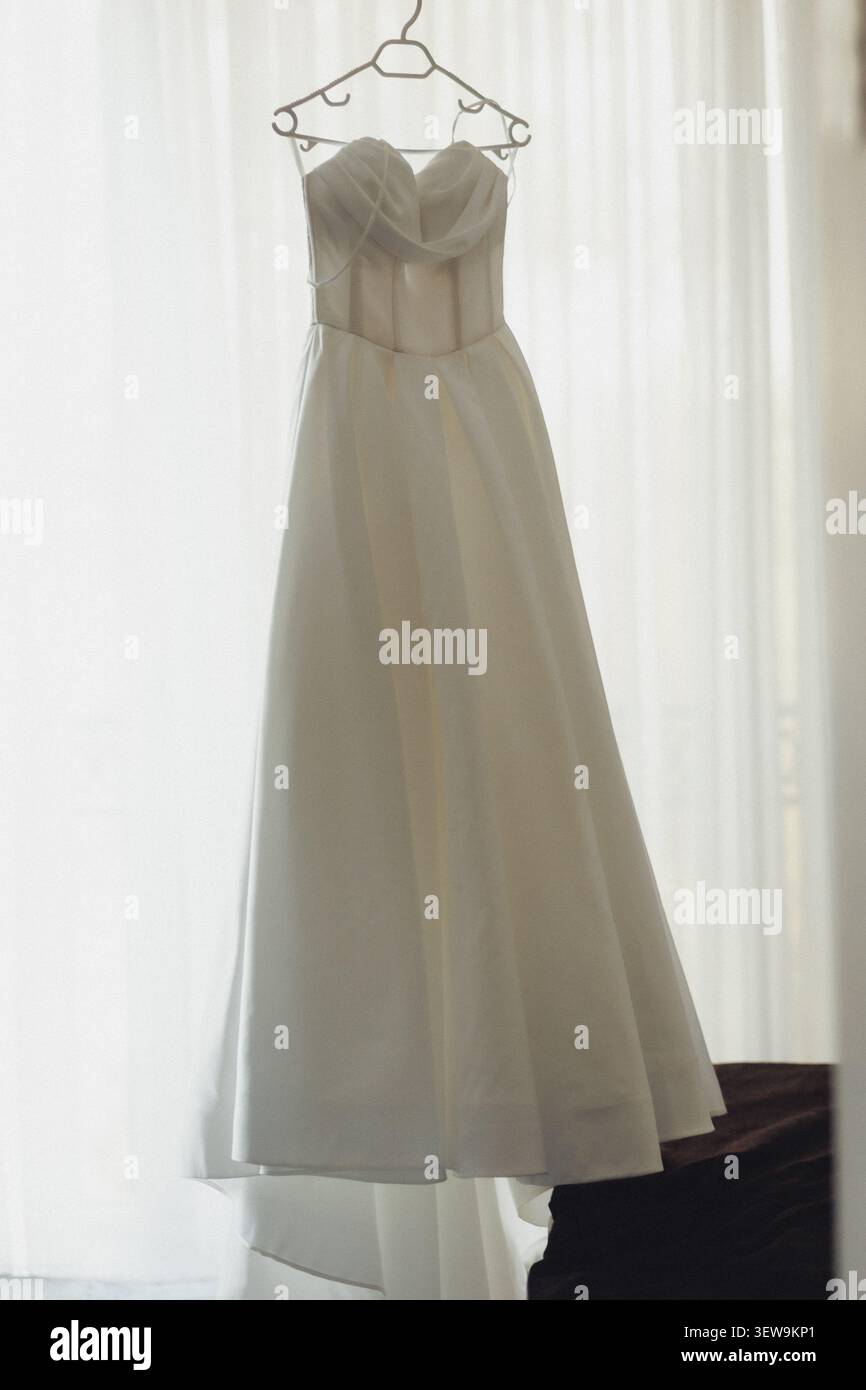 L'elegante abito da sposa è appeso a un gancio in filo metallico. La luce soffusa filtra attraverso le tende trasparenti dietro. Il tessuto si avvolge con grazia, creando pieghe delicate. Silen Foto Stock