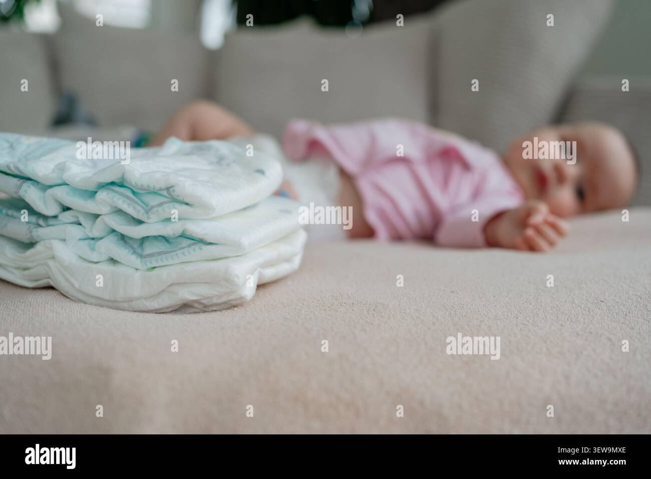 Una pila ordinata di pannolini bianchi usa e getta in primo piano con una bambina appena nata sfocata sdraiata sullo sfondo su un comodo divano. Assistenza all'infanzia e.. Foto Stock