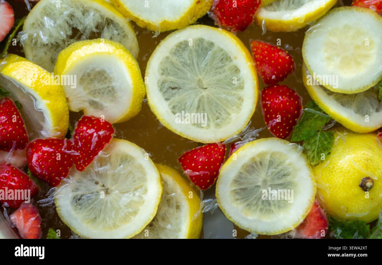 Fette di limone fluttuanti e fragole con foglie di menta in liquido raffreddato creano una rinfrescante estate luminosa, un contrasto di colori vivace, una texture lucida e uno stile alimentare fresco visto dall'alto Foto Stock