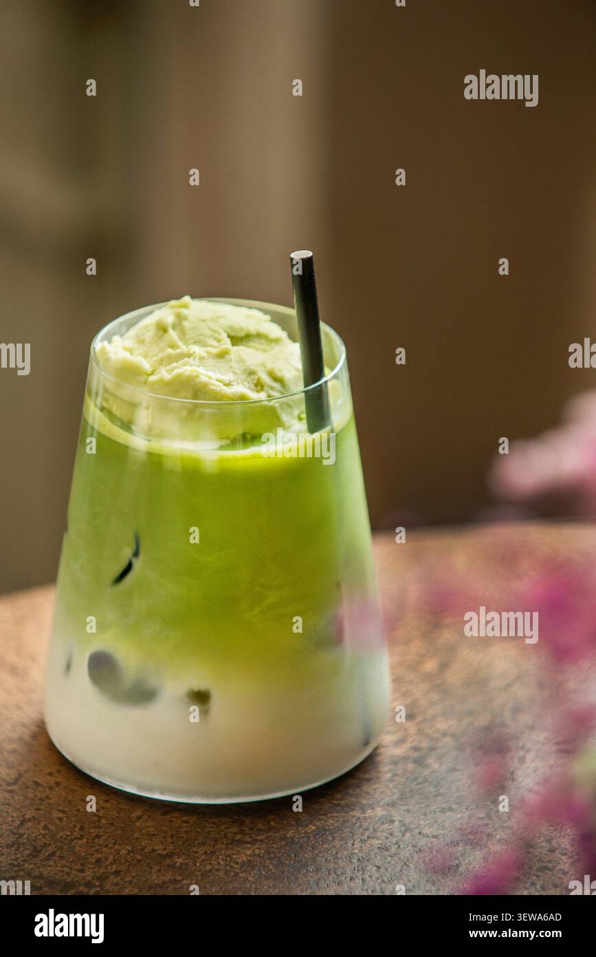 Latte matcha freddo con gelato al tè verde e paglia nera, con bokeh floreale rosa in primo piano. Foto Stock