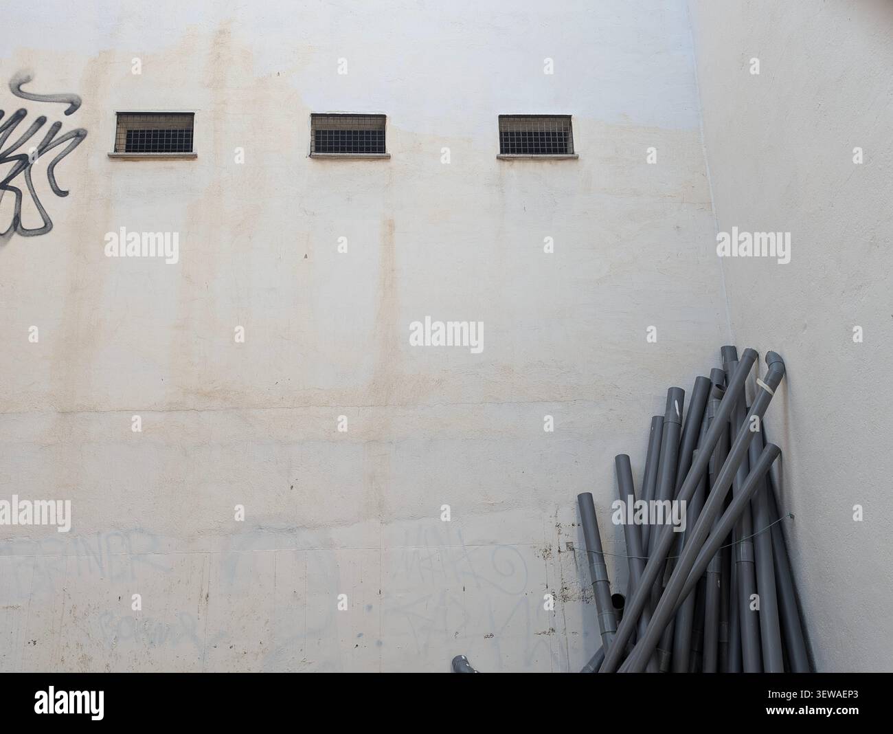 Un mucchio di tubi in PVC grigio impilati nell'angolo di una parete in cemento bianco intemprato con piccole finestre a griglia e graffiti sbiaditi. - Immagine stock catturata con smartphone