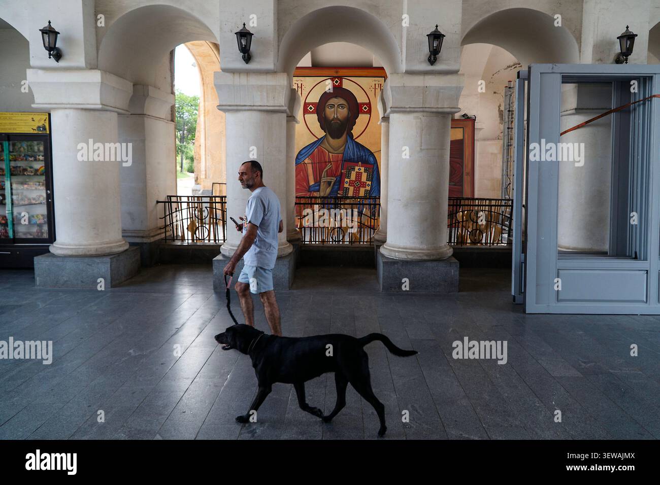 Un uomo cammina un cane nero attraverso una chiesa con colonne e icone sullo sfondo. Foto Stock