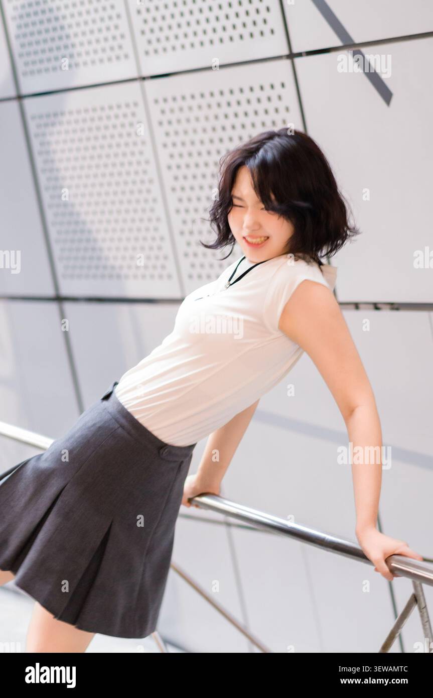 Una giovane donna asiatica allegra con una maglietta bianca e una gonna grigia sorride e si appoggia all'indietro su una ringhiera metallica al chiuso. Foto Stock