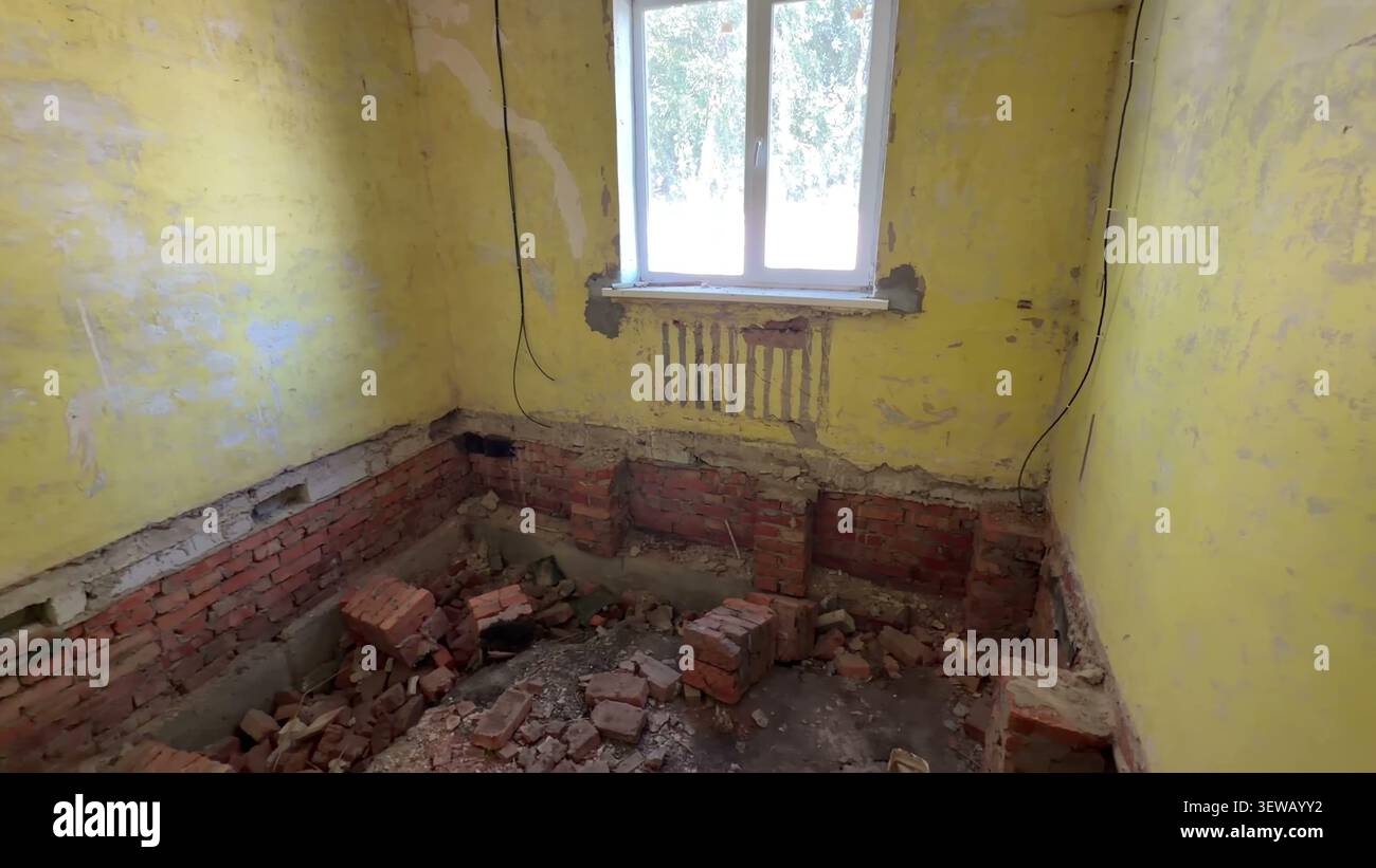 Una sala interna in fase di ristrutturazione presenta pareti gialle con vernice sbucciata, una finestra e fondamenta in mattoni a vista con macerie sul pavimento. Foto Stock