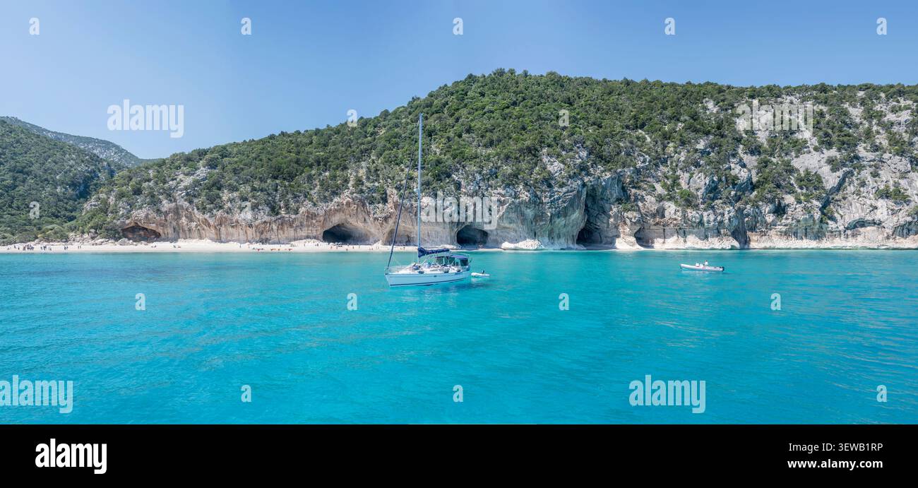 CALA GONONE, 2026 aprile 26; grande paesaggio panoramico editoriale con barca a vela ormeggiata, caverne avvallamenti e gente sparsa sulla spiaggia di Cala Luna On Foto Stock